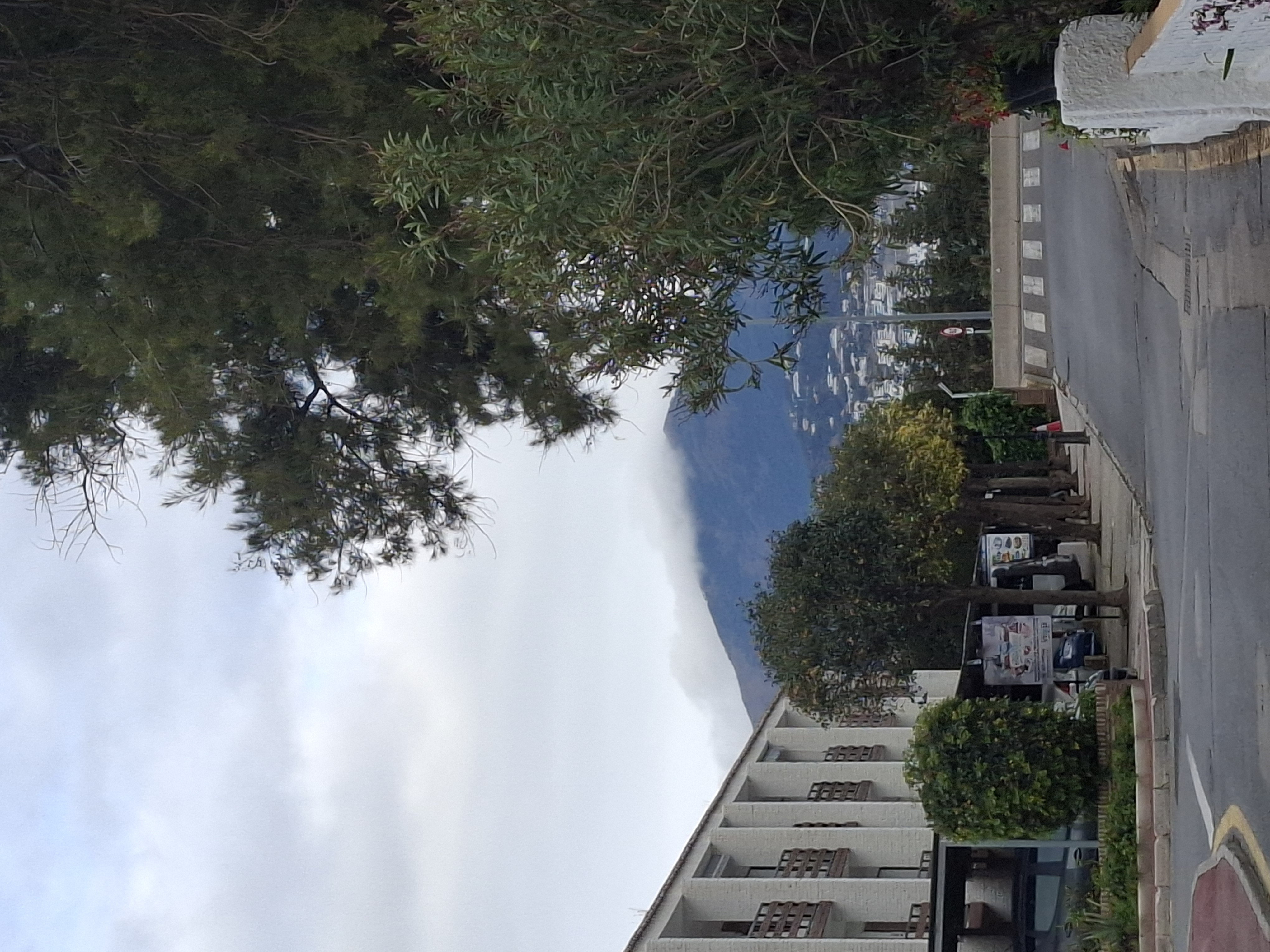 A photo of a street with a dark green mountain in the landscape. It's possible to see a set of white buildings on the left, a mountain with some grayish clouds on its top in the center, and a path of trees on the right. The street is asphalt-colored. A bit far from the point of view, you can see some street and commercial signs, but they are unreadable. There are no visible persons in the photo.