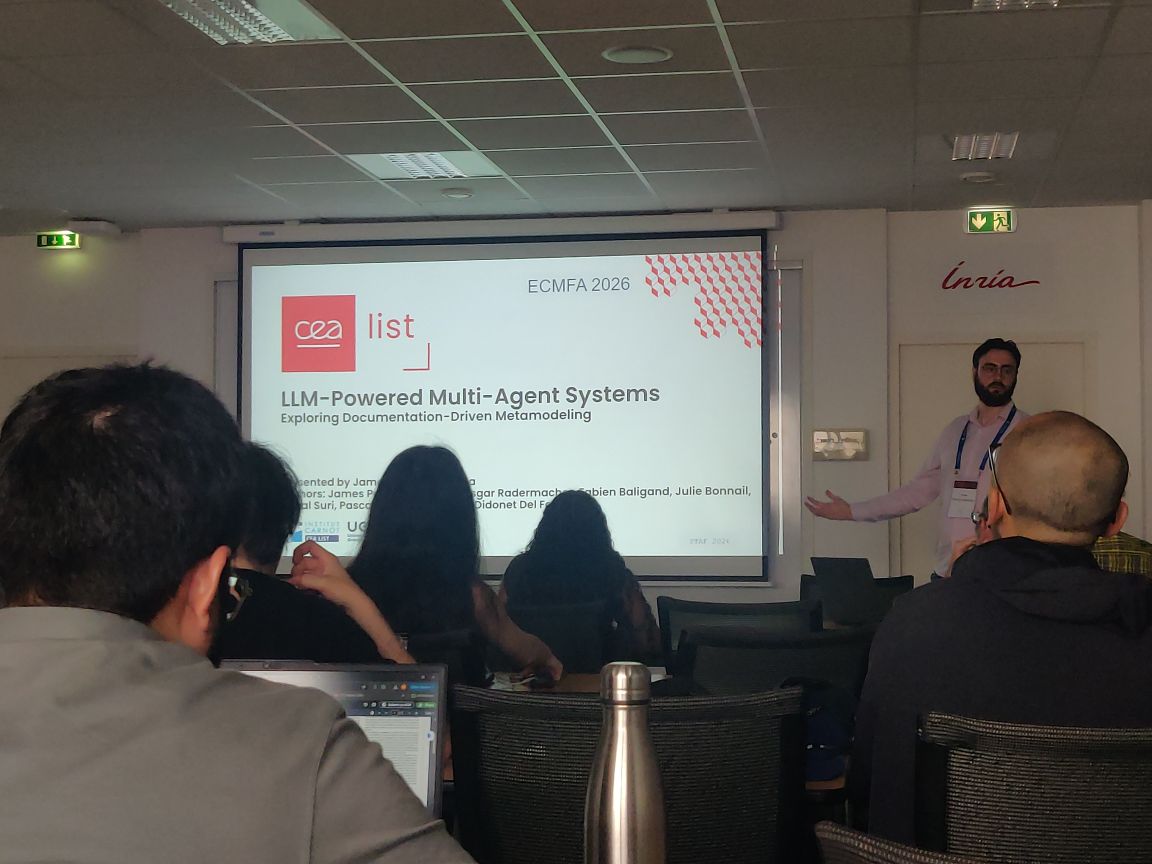  View from the audience of a conference room, where a bearded presenter stands beside a projected slide titled “LLM-Powered Multi-Agent Systems: Exploring Documentation-Driven Metamodeling” at ECMFA 2026. Attendees are seated in the foreground. It's possible to identify 5 persons seated